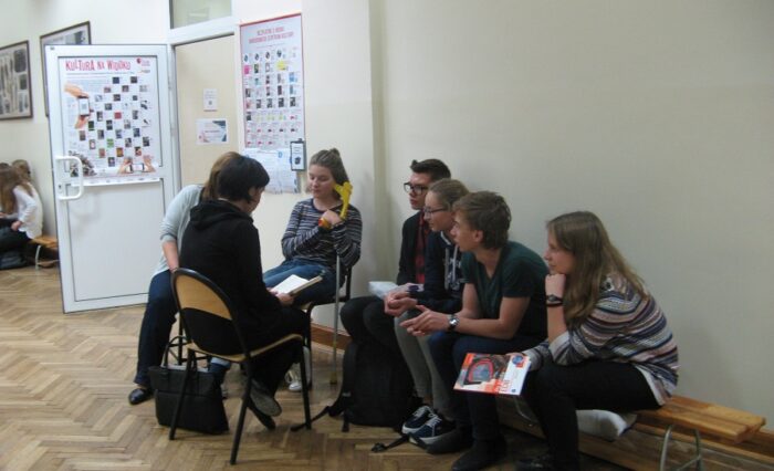 Na zdjęciu grupka młodzieży na ławce przed biblioteką słucha czytającej nauczycielki.