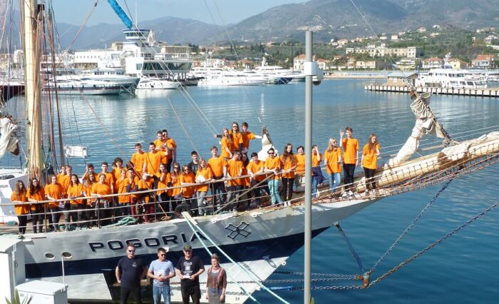 Na zdjęciu grupa młodzieży w pomarańczowych koszulkach na dziobie żaglowca Pogoria.