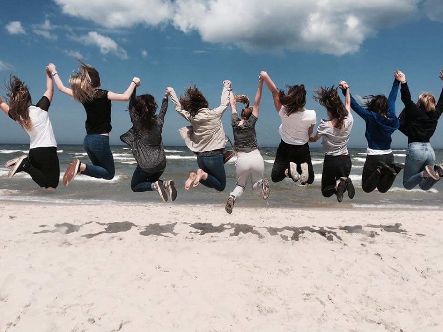 Na zdjęciu grupka młodzieży tyłem skacze na piasku na tle morza.
