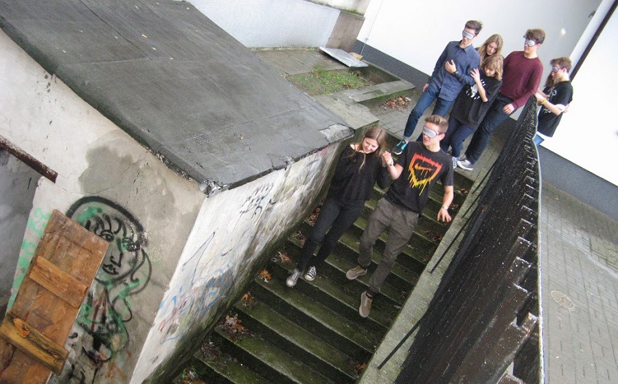Na zdjęciu grupa młodzieży, część z nich z zakrytymi oczami, schodzi po schodach do piwnicy.