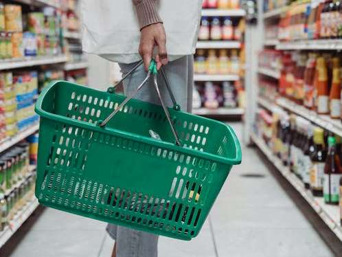 Na zdjęciu ręka trzymająca kosz na zakupy, w tle półki supermarketu.