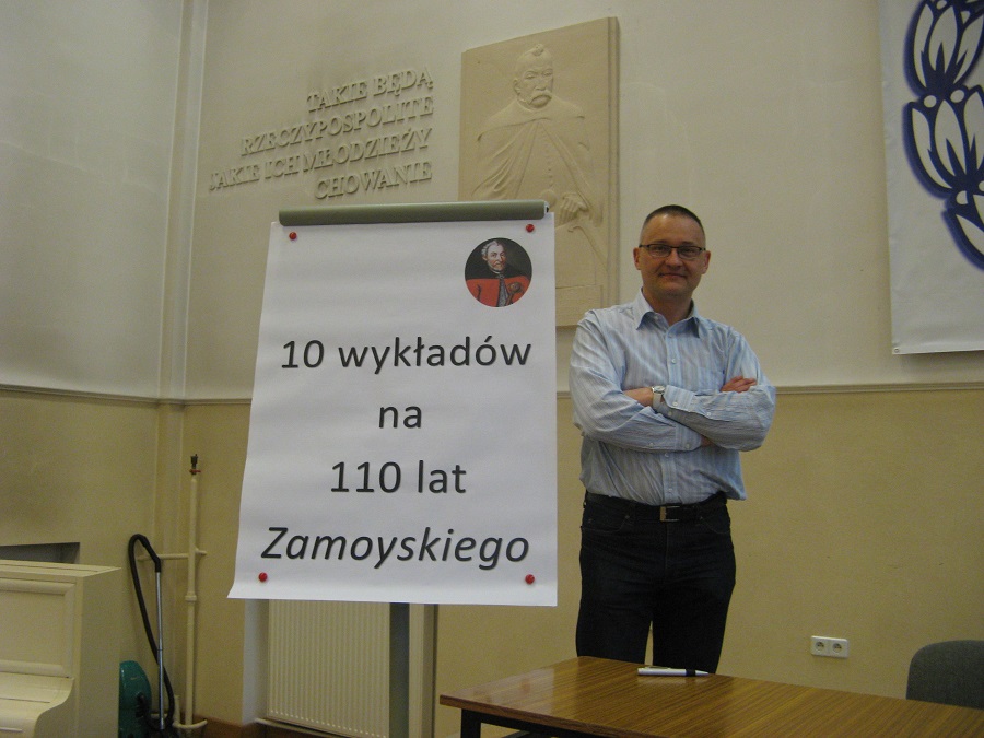 Na zdjęciu wykładowca stoi przy tablicy z napisem "10 wykładów na 110 lat Zamoyskiego".