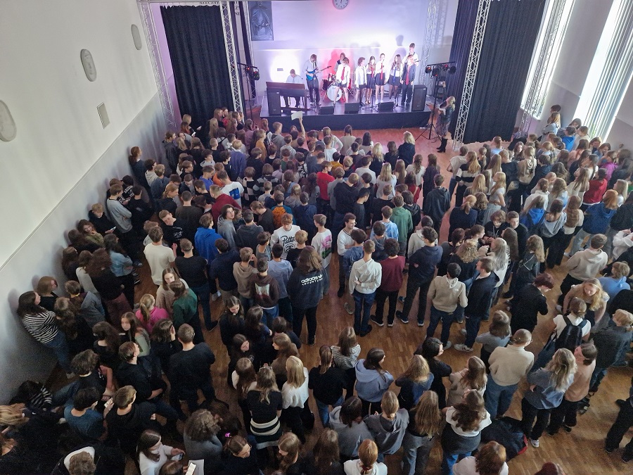 Na zdjęciu artyści na scenie i społeczność szkolna zgromadzona w auli na koncercie.