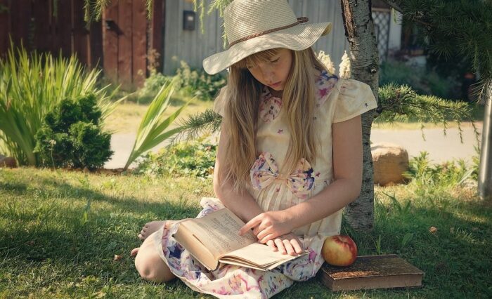 Na zdjęciu dziewczynka w kapeluszu czytająca książkę w ogrodzie.