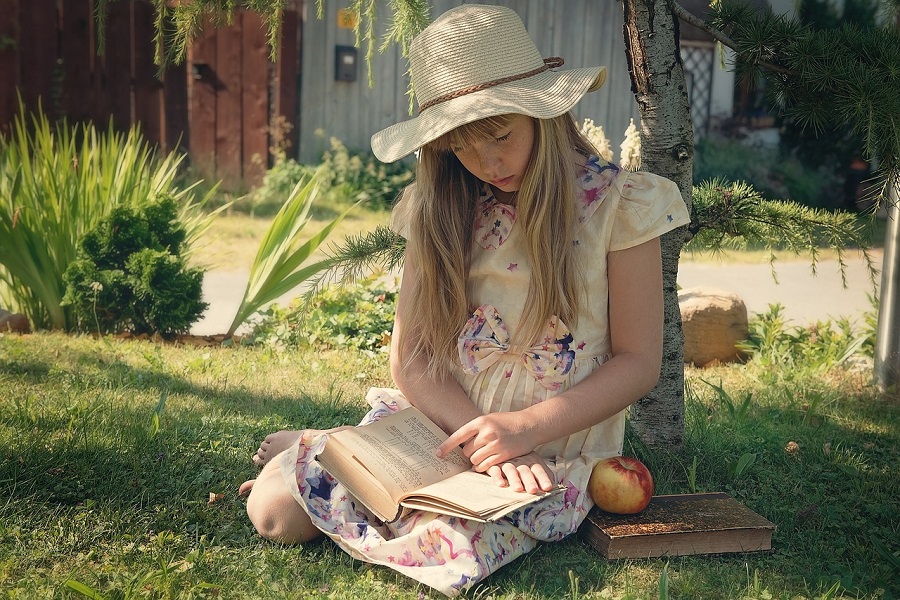 Na zdjęciu dziewczynka w kapeluszu czytająca książkę w ogrodzie.